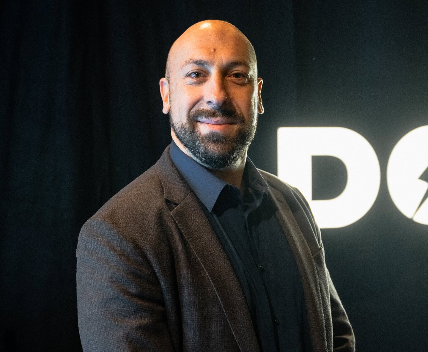 Michael Waksman A man with a beard wearing a dark suit and shirt stands against a black background with partially visible white lettering. He has a bald head and is smiling while looking at the camera.